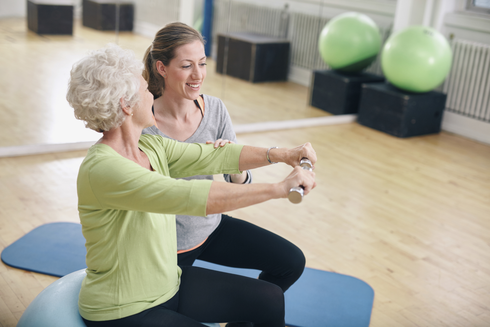 A smiling physical therapist kneeling beside a senior woman who is sitting on an exercise ball holding weights in each hand with her arms extended into a front lateral raise.