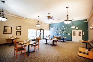 A large dining room with a large window and door. Several tables are placed throughout the room and are set with placemats, cloth napkins and silverware. Five round chandeliers hang from the ceiling.