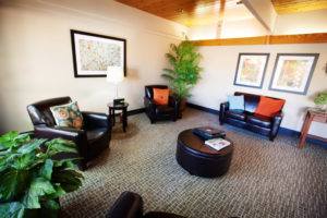 A large waiting area with a leather love seat, three leather chairs, an ottoman, a side table with a lamp, and colorful painting on the walls.